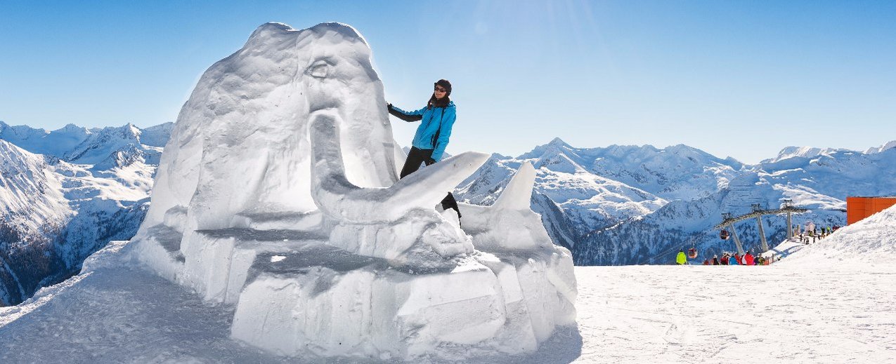 Schneeskulptur Mammut mit Schifahrerin im Vordergrund, Hintergrund blauer Himmel und Berge