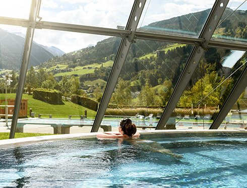 Frau entspannt im Innenbecken der Alpentherme Gastein und blickt durch ein großes Panoramafenster auf die herbstliche Landschaft.