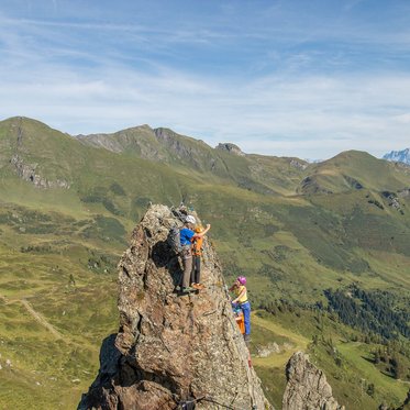 Die Szene zeigt zwei Kletterer, die in einer atemberaubenden Berglandschaft an einem Felsen klettern, umgeben von Natur.