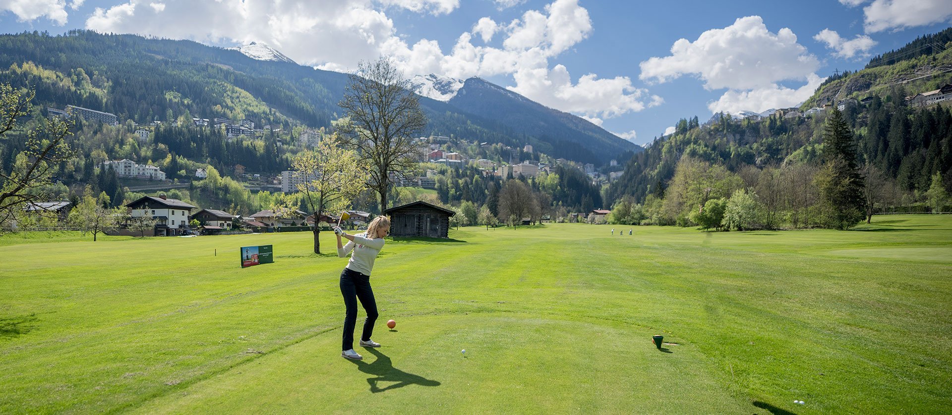 Eine Golferin bereitet sich auf den Abschlag auf einem grünen Golfplatz mit Bergen im Hintergrund vor.