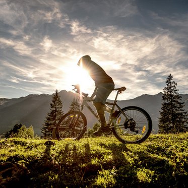 Mountainbiker unterwegs in den Bergen. Die Sonne geht im Hintergrund unter, der Biker wirft einen Schatten. Er ist auf einem Trail in Gastein unterwegs.