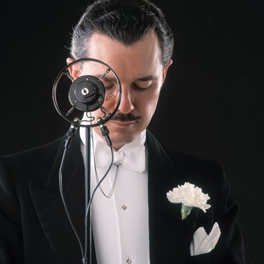 Mann im Smoking und mit Monokel vor einem großen Mikrofon