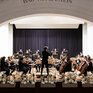 Ein Orchester mit vielen Musikern spielt auf einer Bühne, während der Dirigent leidenschaftlich das Stück leitet.