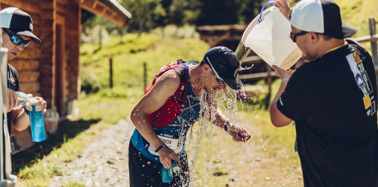 Trailrunner wird bei einer Verpflegungsstation während eines Berglaufs in den Alpen mit Wasser abgekühlt, während Helfer ihn unterstützen.