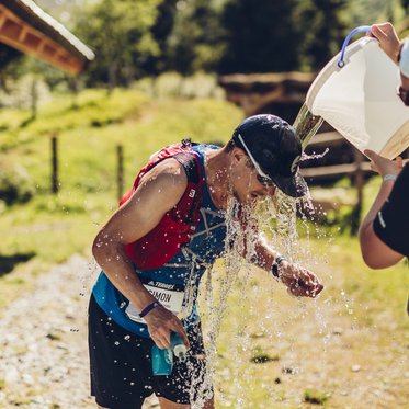 Trailrunner wird bei einer Verpflegungsstation während eines Berglaufs in den Alpen mit Wasser abgekühlt, während Helfer ihn unterstützen.