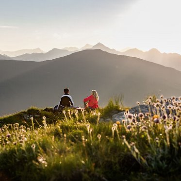 Zwei Personen sitzen auf einem Hügel und genießen den Sonnenuntergang über den Bergen, umgeben von blühenden Pflanzen.